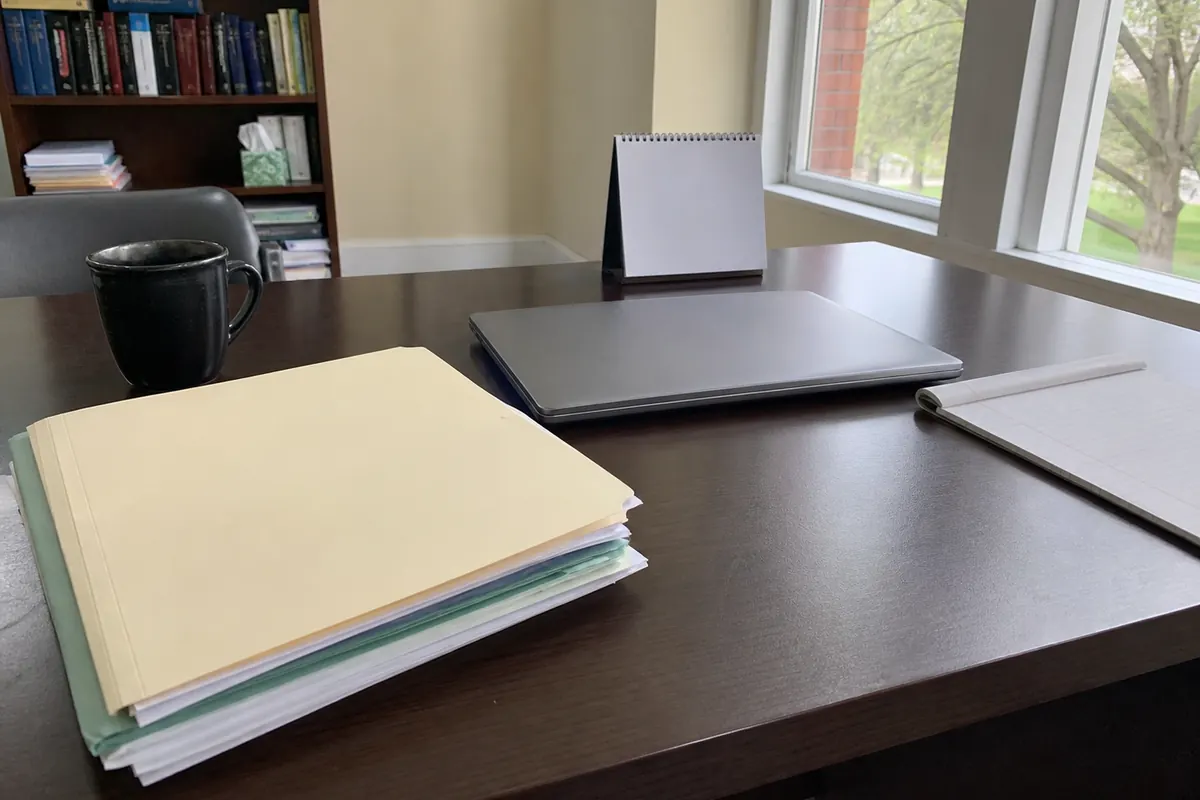 A thick stack of blank manila application folders next to a closed laptop and a small flip desk calendar, a dark coffee mug, and the edge of a spiral notepad, on a wood office desk with a bookshelf and a window looking out on trees.