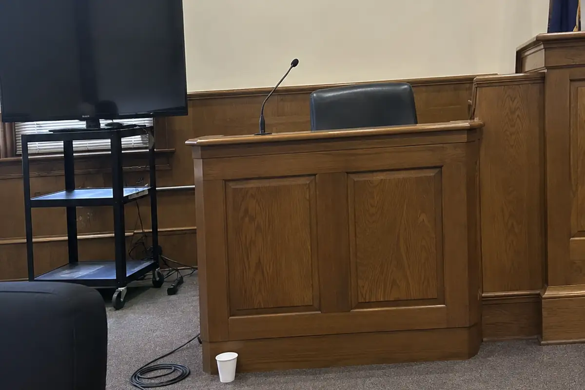 An empty wooden witness stand with a black chair and a short gooseneck microphone in a small wood-paneled courtroom, a blank dark flat screen on a rolling cart beside it, a coiled cable and a paper cup left on the carpet, in ordinary flat daylight.