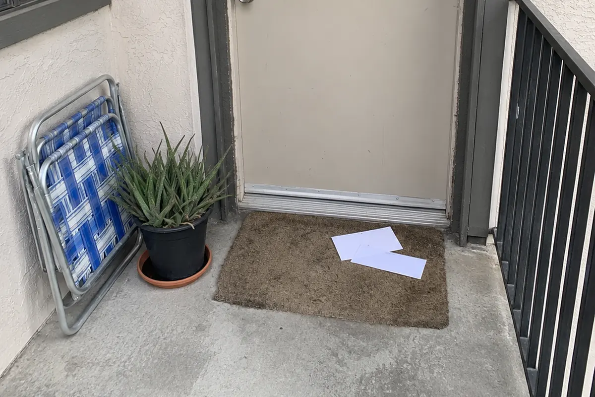 Blank white papers left on a worn doormat outside the front door of an apartment unit, with a folded lawn chair and a potted aloe beside the doorway and an outdoor walkway railing at the edge of the frame, in flat overcast daylight.