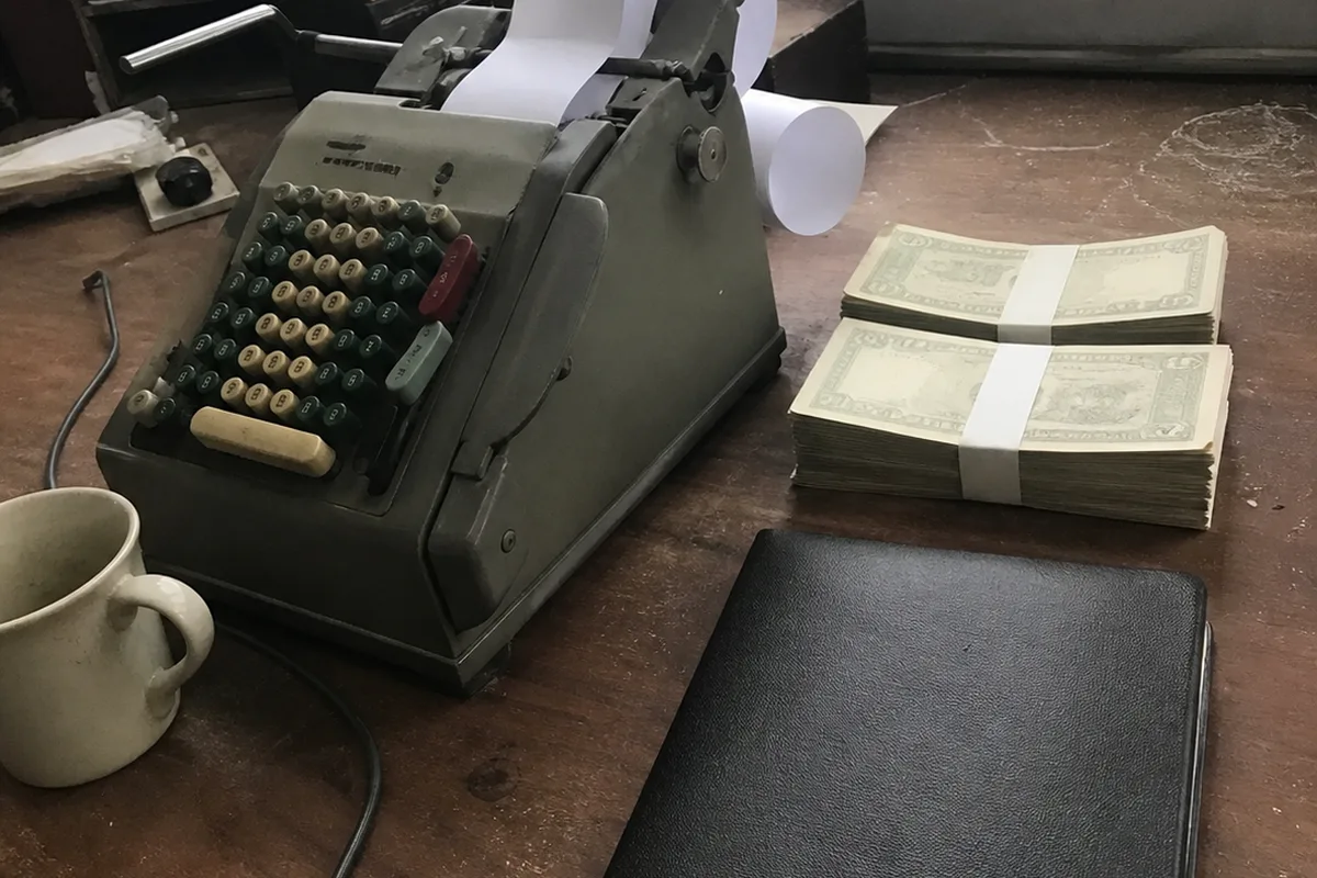A blank checkbook, a mechanical adding machine, and a closed leather ledger stacked on a wooden desk in a law office, soft morning daylight through venetian blinds casting striped shadows, no legible text anywhere.