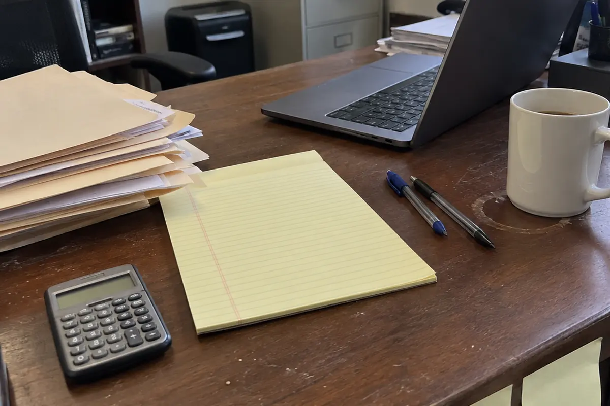 A scuffed wooden desk in a small office under flat daylight: a black pocket calculator with a blank display at the near corner, an unwritten yellow legal pad in the middle, two ballpoint pens beside it, a white coffee mug sitting on a faint ring stain, a leaning stack of manila folders and loose paper on the left, and an open laptop angled away so its screen cannot be seen.