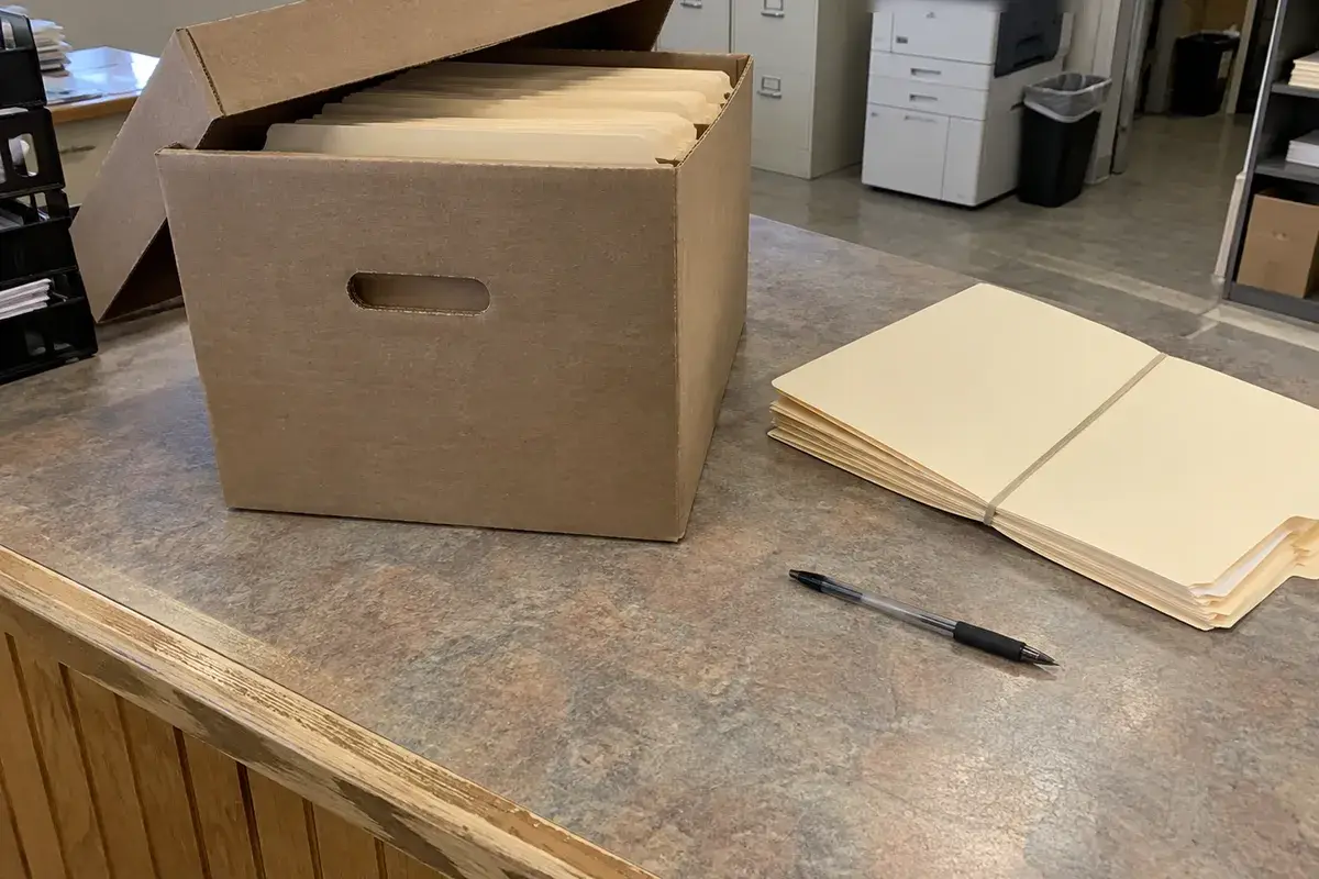A plain cardboard records box with its lid pushed back sits on a worn laminate service counter in a records office, filled with unlabeled manila folders, beside a rubber-banded bundle of blank folders and a pen. Filing cabinets stand in the background. No people are in the frame.