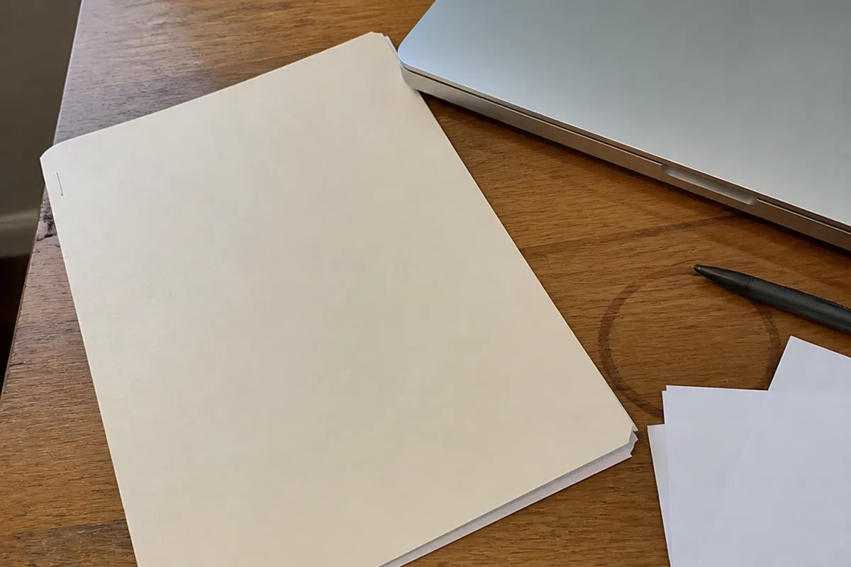 A stapled case folder set down on a cluttered desk next to a closed laptop, a coffee ring on the wood, a pen, and loose papers turned face-down, ordinary daylight from a window.