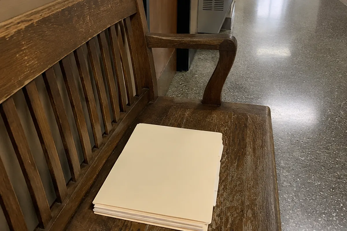 A stack of blank manila folders left on a worn wooden courthouse hallway bench, a water fountain and an open doorway in the background, ordinary hallway daylight and fluorescent light.