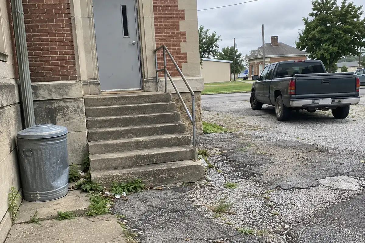 Worn concrete steps and a plain metal handrail at the side door of a small brick county courthouse, a dented galvanized trash can beside them, weeds growing through cracked asphalt and one older pickup truck parked on the gravel lot, in flat overcast daylight.
