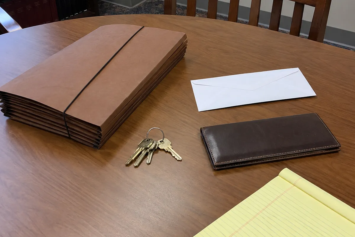 A round wooden conference table in flat office daylight: a closed brown accordion file held shut by an elastic band, a ring of brass house keys, a blank white envelope, a closed dark leather checkbook cover and the corner of a blank yellow legal pad, with an empty wooden chair pushed in behind. No people are in the frame.