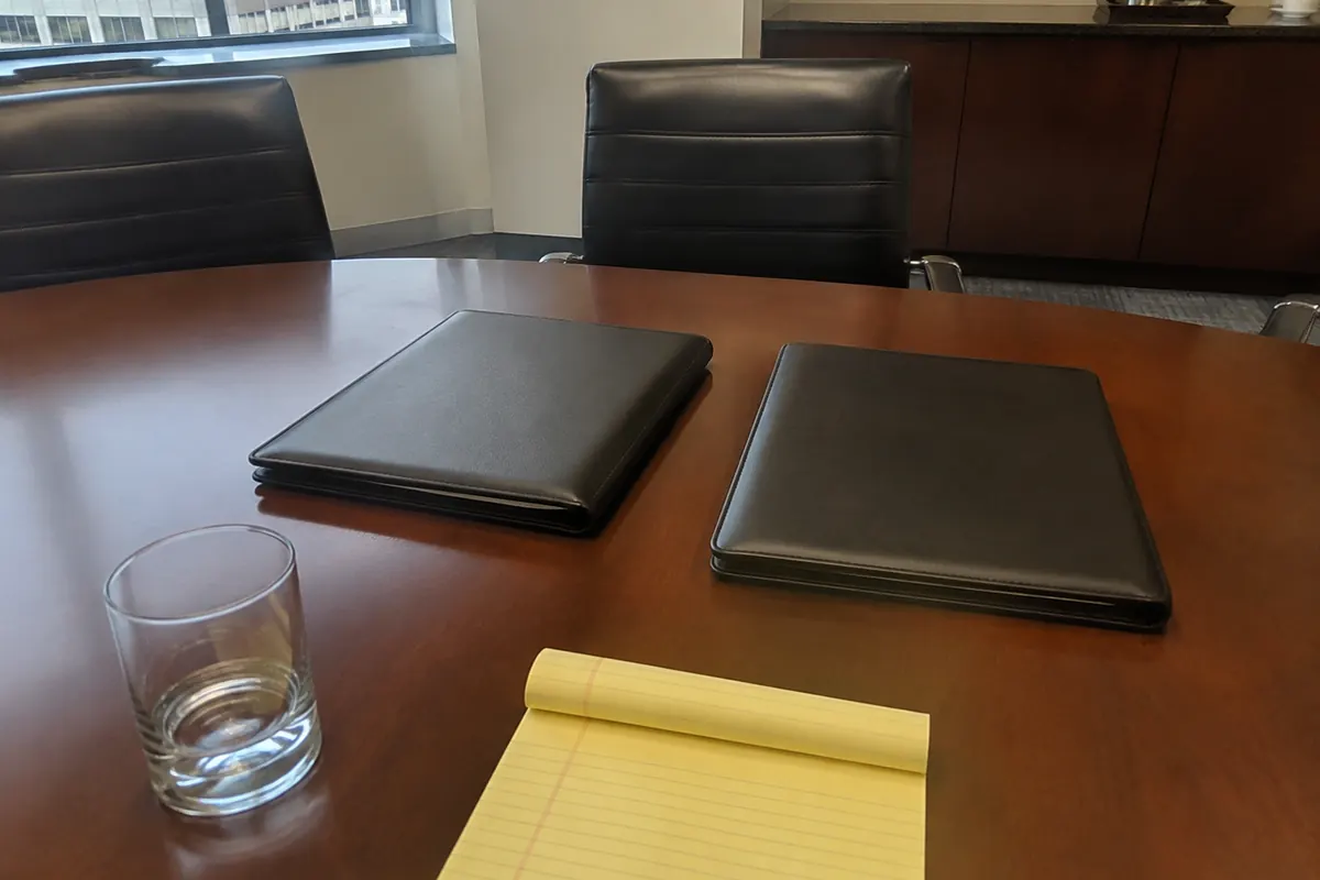 Two closed black leather portfolios on a law firm conference table, a water glass and a yellow legal pad nearby, empty leather chairs and a credenza in the background, ordinary daylight from a city window.