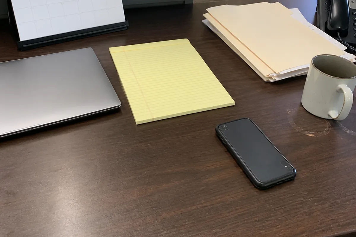 A yellow legal pad, a closed laptop, a stack of manila folders, and a phone face-down on a law office desk, with a desk calendar and a black telephone in the background, ordinary daylight from a window.
