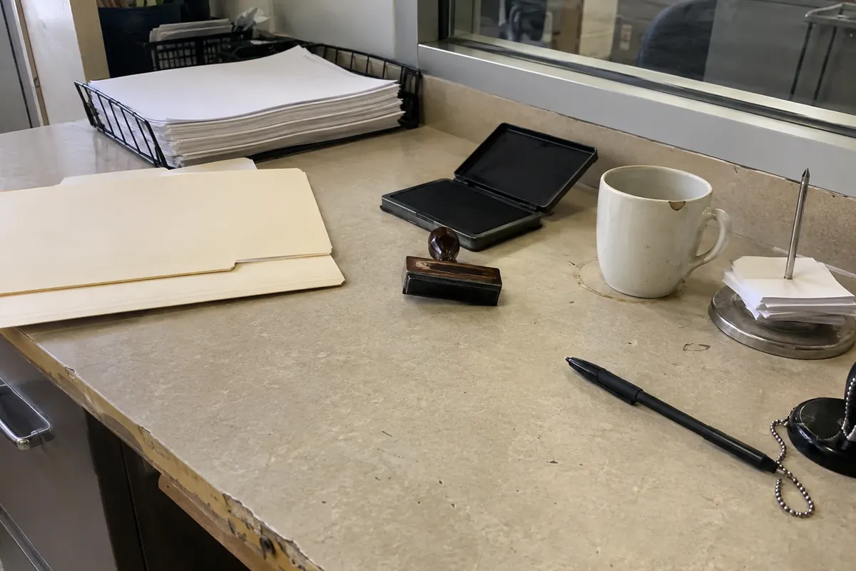 The corner of a worn beige laminate counter at a courthouse clerk's window in flat daylight: a black wire document tray holding a thick stack of blank white paper, two closed manila folders with blank tabs, a wooden-handled rubber date stamp resting face down beside an open black ink pad, a chipped white mug on a faint ring stain, a metal spindle of blank slips and a ballpoint pen on a beaded chain.