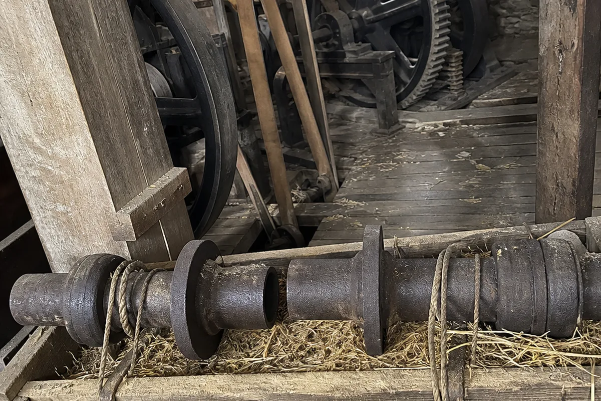 A broken iron crankshaft resting on a wooden crate inside a stilled flour mill, mill gears and leather drive belts hanging motionless in the background, dust motes visible in one warm amber light against deep green shadow.