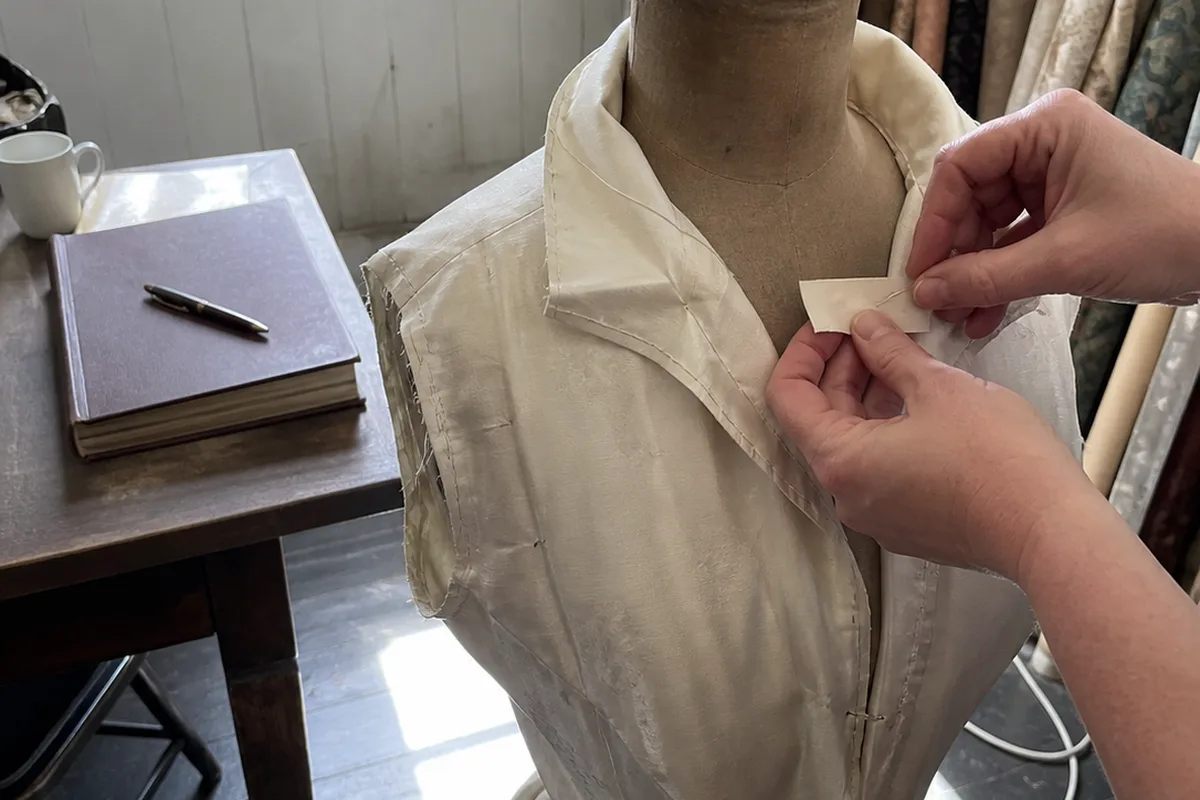A dress form draped in fine fabric in an early 20th-century fashion atelier, bolts of fabric leaning against the wall, a blank exclusivity contract and a fountain pen on a wooden table, golden-hour sunlight through tall windows.