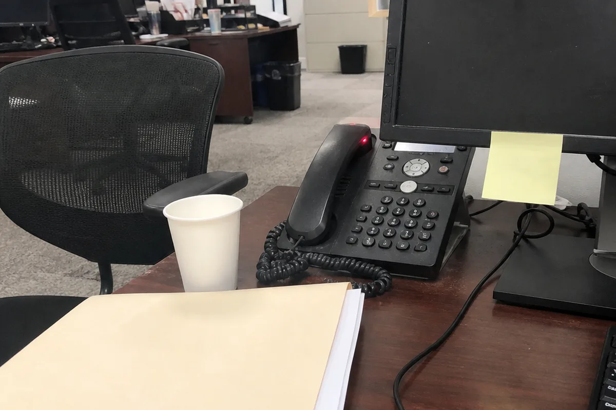 A desk phone with its hold button lit red, beside a closed manila case folder and a yellow sticky note curling at the corner tucked under a dark computer monitor, an empty office chair pushed back in the background, one warm desk lamp against deep green shadow.