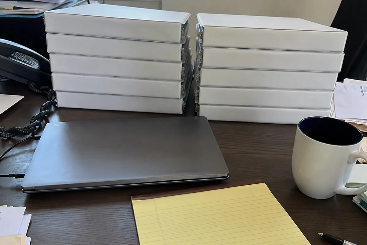 Two nearly identical stacks of blank white binders side by side on a law office desk, next to a closed laptop, a yellow legal pad, and a coffee mug, ordinary daylight from a city window.