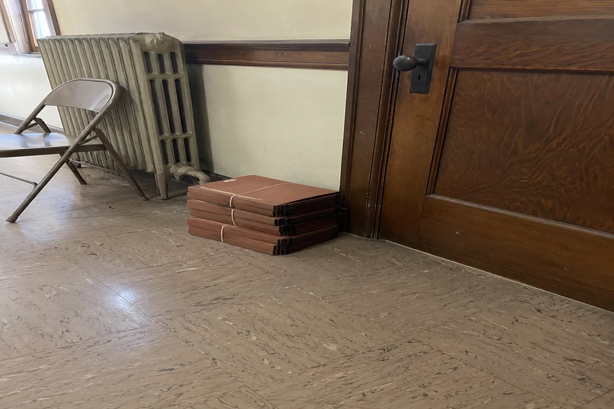A stack of worn accordion case folders bound with rubber bands on the floor beside a heavy wooden courtroom door in a plain courthouse hallway, a cast iron radiator and folding chair nearby, flat overcast daylight.