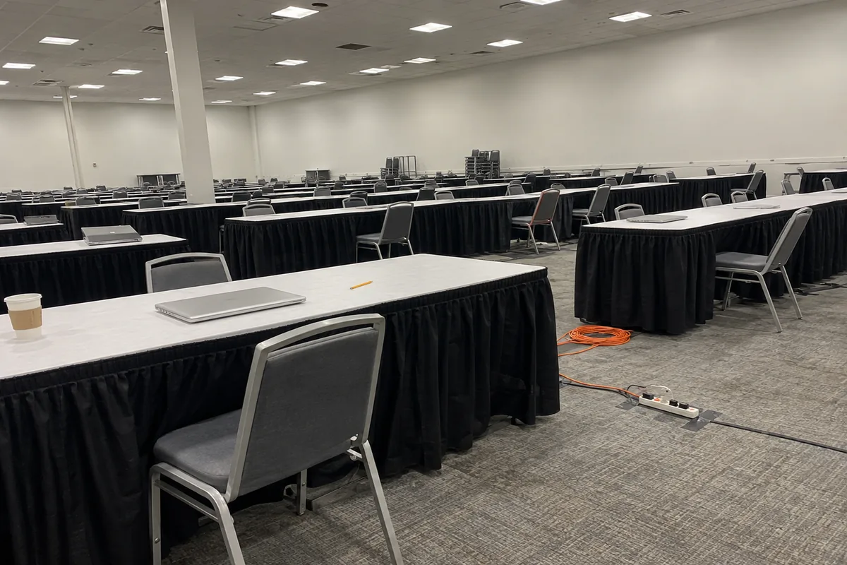 An emptied convention hall set up for a written examination in flat fluorescent daylight: long rows of folding tables under plain black skirting, grey stacking chairs pushed in at uneven angles, two closed silver laptops, a paper cup and a pencil left behind, and a coiled orange extension cord running to a power strip taped down on the grey carpet.