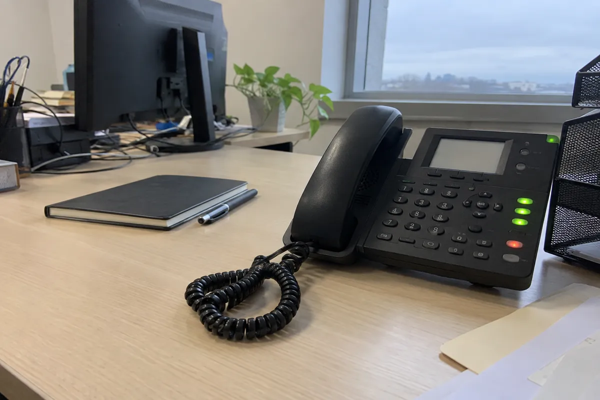 Un teléfono de escritorio de varias líneas sobre un escritorio de madera clara, fotografiado descentrado con luz de día plana y nublada desde una ventana, con el auricular con cable descansando en su base, un cable enroscado sobre el escritorio, varias luces indicadoras pequeñas a lo largo de la base del teléfono, un cuaderno liso cerrado y un bolígrafo al lado, y un monitor de computadora desenfocado y una pequeña planta en maceta al fondo. No hay personas en la imagen.