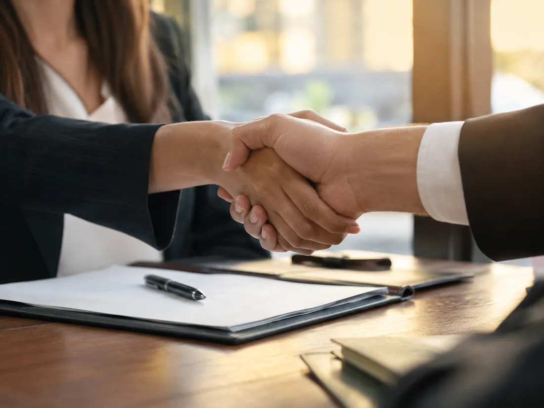 Two people in business attire shaking hands over a desk.